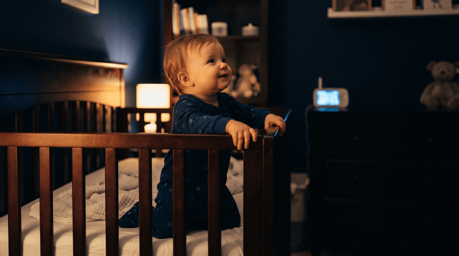 An 8-month-old baby pulling up to stand at the edge of a dark nursery crib at night